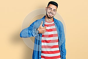 Young hispanic man with beard wearing casual denim jacket doing happy thumbs up gesture with hand