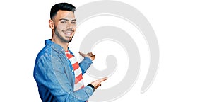 Young hispanic man with beard wearing casual denim jacket inviting to enter smiling natural with open hand