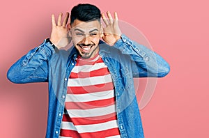 Young hispanic man with beard wearing casual denim jacket trying to hear both hands on ear gesture, curious for gossip