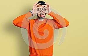 Young hispanic man with beard wearing casual orange sweater doing ok gesture like binoculars sticking tongue out, eyes looking