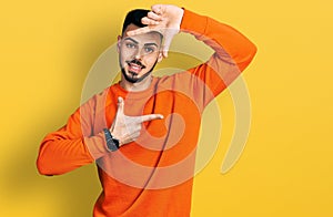 Young hispanic man with beard wearing casual orange sweater smiling making frame with hands and fingers with happy face