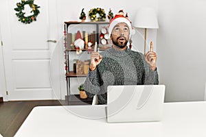Young hispanic man with beard wearing christmas hat using laptop amazed and surprised looking up and pointing with fingers and