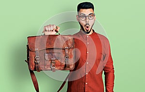 Young hispanic man with beard wearing leather bag scared and amazed with open mouth for surprise, disbelief face