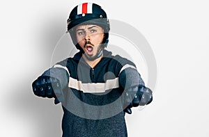 Young hispanic man with beard wearing motorcycle helmet doing driving gesture in shock face, looking skeptical and sarcastic,