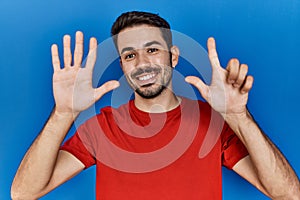 Young hispanic man with beard wearing red t shirt over blue background showing and pointing up with fingers number seven while