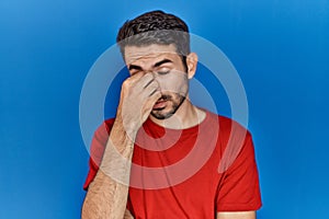 Young hispanic man with beard wearing red t shirt over blue background tired rubbing nose and eyes feeling fatigue and headache