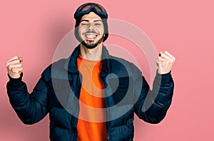 Young hispanic man with beard wearing snow wear and sky glasses celebrating surprised and amazed for success with arms raised and