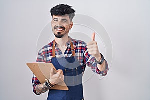 Young hispanic man with beard wearing waiter apron holding clipboard approving doing positive gesture with hand, thumbs up smiling