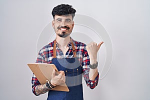 Young hispanic man with beard wearing waiter apron holding clipboard pointing to the back behind with hand and thumbs up, smiling