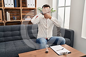 Young hispanic man with beard working at consultation office covering eyes and mouth with hands, surprised and shocked