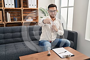 Young hispanic man with beard working at consultation office punching fist to fight, aggressive and angry attack, threat and