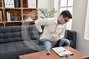 Young hispanic man with beard working at consultation office suffering of backache, touching back with hand, muscular pain