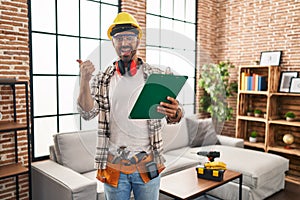 Young hispanic man with beard working at home renovation pointing thumb up to the side smiling happy with open mouth