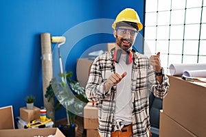 Young hispanic man with beard working at home renovation pointing to the back behind with hand and thumbs up, smiling confident