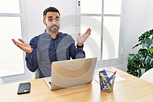 Young hispanic man with beard working at the office with laptop clueless and confused expression with arms and hands raised