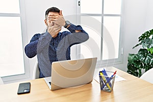 Young hispanic man with beard working at the office with laptop covering eyes and mouth with hands, surprised and shocked