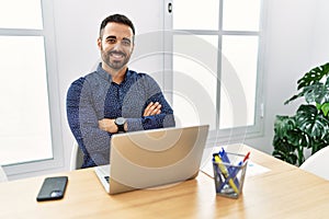 Young hispanic man with beard working at the office with laptop happy face smiling with crossed arms looking at the camera