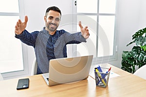 Young hispanic man with beard working at the office with laptop looking at the camera smiling with open arms for hug