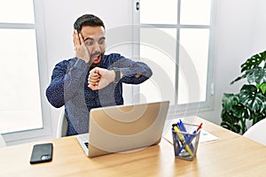 Young hispanic man with beard working at the office with laptop looking at the watch time worried, afraid of getting late