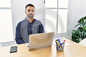 Young hispanic man with beard working at the office with laptop relaxed with serious expression on face