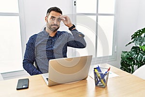 Young hispanic man with beard working at the office with laptop worried and stressed about a problem with hand on forehead,