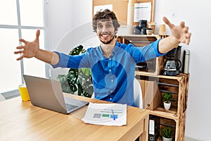 Young hispanic man with beard working at the office using computer laptop looking at the camera smiling with open arms for hug