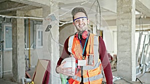 Young hispanic man builder smiling confident holding hardhat at construction site