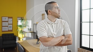 Young hispanic man business worker standing with relaxed expression and arms crossed gesture at office