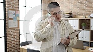 Young hispanic man business worker talking on the phone reading from clipboard at office