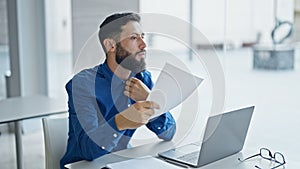 Young hispanic man business worker using document as a handfan at the office