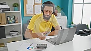 Young hispanic man business worker using headphones and smartphone working at the office
