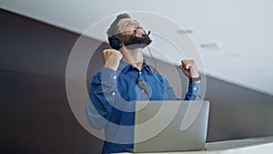 Young hispanic man business worker using laptop and headphones celebrating at the office