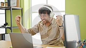 Young hispanic man business worker using laptop and headphones celebrating at office