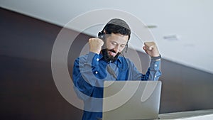 Young hispanic man business worker using laptop and headphones celebrating at the office