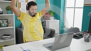 Young hispanic man business worker using laptop and headphones celebrating at the office