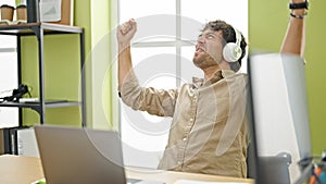 Young hispanic man business worker using laptop and headphones celebrating at office