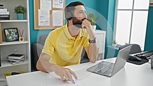 Young hispanic man business worker using laptop and headphones thinking at the office