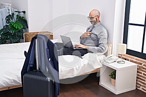 Young hispanic man business worker using laptop sitting on bed at hotel room