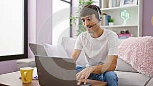 Young hispanic man call center agent using laptop and headphones sitting on sofa at home