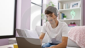 Young hispanic man call center agent using laptop and headphones sitting on sofa at home