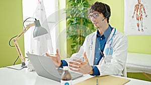 Young hispanic man doctor doing video call with laptop and headphones at the clinic