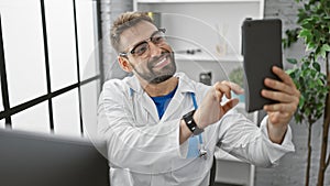 Young hispanic man doctor making selfe picture by touchpad smiling at the clinic