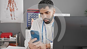 Young hispanic man doctor using smartphone and computer at clinic