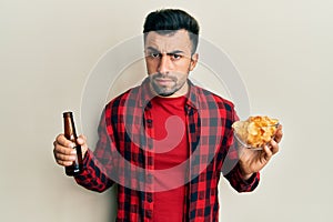 Young hispanic man drinking a bottle of beer and eating chip potatoes skeptic and nervous, frowning upset because of problem