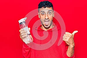 Young hispanic man drinking bottle of water pointing thumb up to the side smiling happy with open mouth