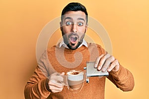 Young hispanic man drinking a cup of coffee with alcohol afraid and shocked with surprise and amazed expression, fear and excited