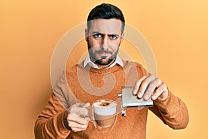 Young hispanic man drinking a cup of coffee with alcohol skeptic and nervous, frowning upset because of problem