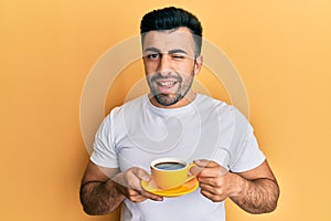 Young hispanic man drinking a cup of coffee winking looking at the camera with sexy expression, cheerful and happy face