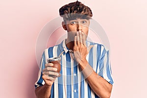 Young hispanic man drinking mate infusion covering mouth with hand, shocked and afraid for mistake