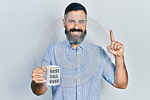 Young hispanic man drinking mug of coffee with best dad ever message smiling with an idea or question pointing finger with happy